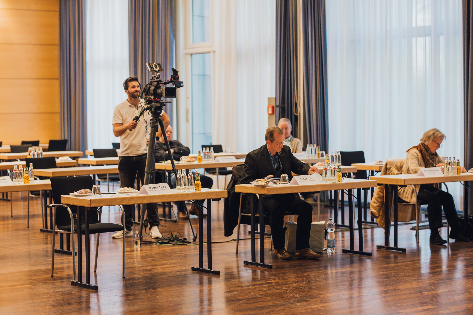 Mitglieder des Runden Tisches an Konferenztischen, links ien Kameramann hinter einer Kamera